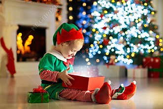 Child opening present at Christmas tree at home. Kid in elf costume with Xmas gifts and toys. Little baby boy with gift box and