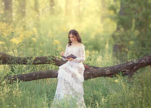 Charming sweet girl with dark hair and bare shoulders in a gorgeous vintage white dress sits on a fallen tree and reads