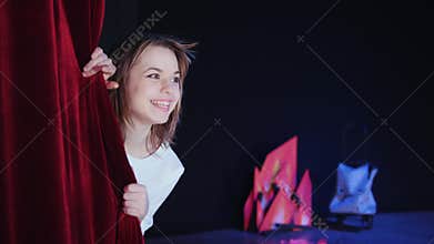 A theater stage. A girl looking out of the backstage cloth, smiles and goes back.