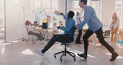 Slow motion crazy fun young black businessman riding office chair, celebrating success with team throwing paper in air.