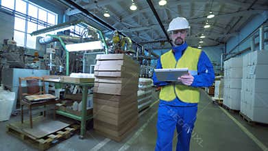 Male worker walks in a facility, typing on a tablet, factory equipment.
