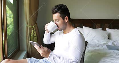 Young Man Using Tablet Computer Drink Coffee Smiling Guy Chatting Online Over Big Window In Bedroom