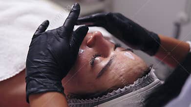 Closeup view of a woman with blemish skin lying on the couch while professional cosmetologist is applying face cream and