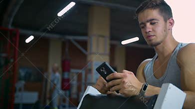 Athletic man sitting in fitness gym after workout, chatting online smart phone