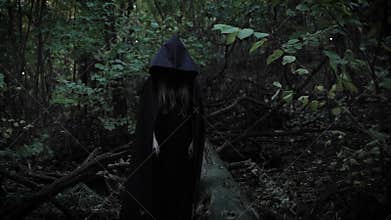 A girl in a black cloak with a hood shows her finger straight, a mystical lady is in a scary park in the evening