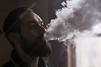 Man with Beard and Mustages Vaping an Electronic Cigarette. Vaper Hipster Smoke Vaporizer and Exhals Smoke Cloud.