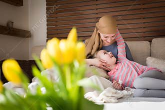 Young adult female cancer patient spending time with her daughter at home, relaxing on the couch. Cancer and family support.