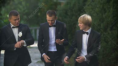 Wedding concept. The young handsome groom is happily talking with his three best man while walking along the garden.