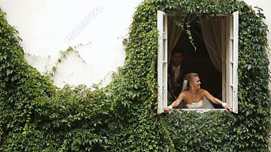 The cheerful bride is opening the window while groom is kissing her. The house is covered with beautiful herbs.