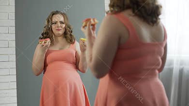 Depressed overweight lady chewing donuts in front of mirror, eating disorder