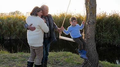 Happy married couple with grandson on swing is having fun near river in sunshine in warm weather