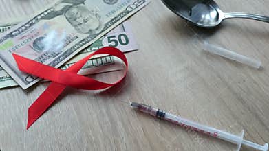 HIV/AIDS concept. Red awareness ribbon dropping on a table with syringe with drugs.