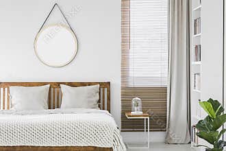 Window with wooden blinds and light grey curtain in white bedroom interior with mockup poster, double bed and rack with books