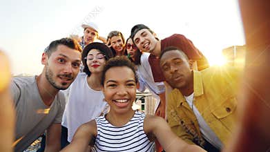 Point of view shot of African American girl holding camera and taking selfie with happy friends at party on roof. Men