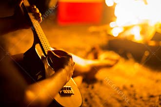 Man strumming an ukulele/guitar in the woods next to an open bonfire