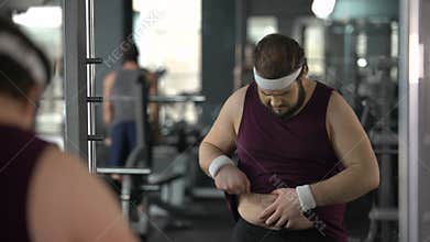 Unhappy overweight man looking at his mirror reflection in gym, diet and sport