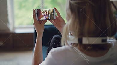 Young woman is talking to friends online with smartphone looking at screen and speaking sitting on couch in modern