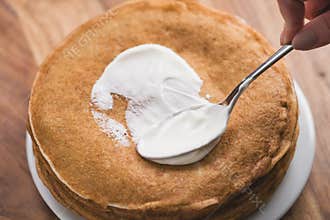 Adiing sour cream to stack of fresh hot crepes or blinis on wood table