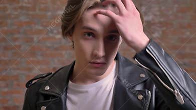 Young attractive man in black leather jacket turning into camera and touching his hair, brick background