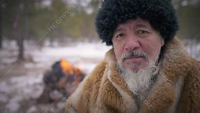 Close-up portrait serious indigenous senior bearded man looking at camera sitting on the right with fire burning at