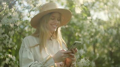 Portrait of Blonde Woman in Hat Enjoy Success on Mobile Phone of White Apple Blossoms. Garden With Blooming Trees. The