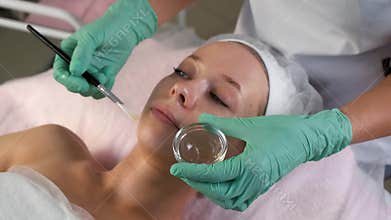 Cosmetologist applying peeling mask on woman face using brush in beauty clinic.