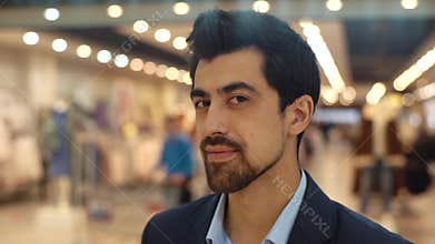 Close-up of confident bearded young man in suit turning head, looking at the camera and smiling standing in modern