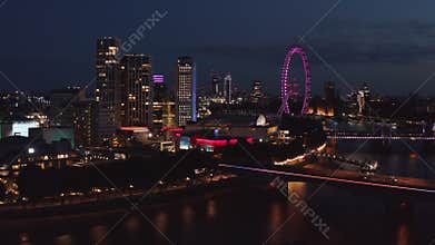 Forwards fly above city at night. Illuminated buildings on Thames river waterfront. Royal Festival Hall and violet