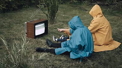 Family are sitting on the grass in raincoats and watching an old retro TV