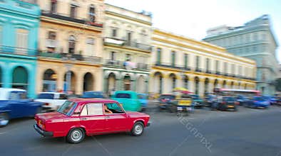 A small iconic red classic old car fast moving speeding fast along Paseo de Marti with colourful colonial buildings, Havana, Cuba