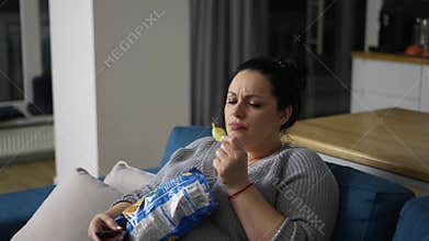 Overweight woman sitting on couch at home, watching tv and eating potato chips