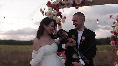 Funny portrait of laughing newlyweds with a dachshund in their arms near a flower arch in the countrysidere