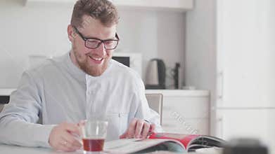 A young stylish man with glasses in the morning at breakfast reads a magazine and drinks tea