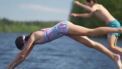 Girl and little boy in wetsuit jumping into the lake from wooden pier. Having fun on summer day.