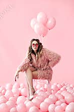 Confident fashion woman sitting on chair posing on many pink balloons background