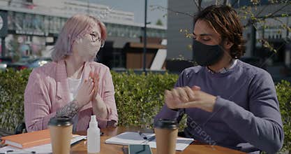Students wearing masks studying in street cafe spraying hands with sanitizer