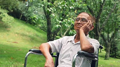 Disabled senior man in eyeglasses sitting in wheelchair looking away and thinking with his hand on chin in the park.