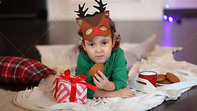 Little girl in funny deer mask and Christmas pajamas lie on a plaid eating cookies and drinking cocoa