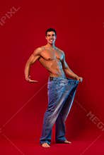 Young man showing how much weight he lost with big jeans. Fat to fit concept. Fitness poster on red background