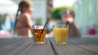 Close-up orange and apple juice on wooden poolside in sunshine with blurred happy children resting at background