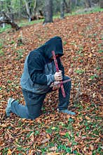 A man in a hood, bowing his head, stands on one knee in prayer and holds a cross below