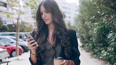 Smiling young business woman using her mobile phone while drinking coffee walking down the street.