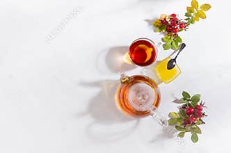 Autumn healthy rosehip tea in transparent teapot with cup, honey, dog rose branch in bright cozy sun beams white wood table top.