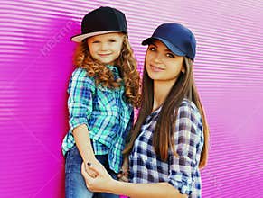 Portrait of young mother with child little girl wearing a checkered shirts and baseball cap in city over pink