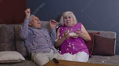 Scared senior old couple holding popcorn watching horror tv show film sitting on sofa in living room