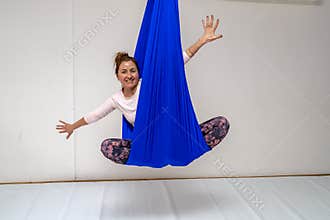 A young woman poses while doing anti-gravity aerial yoga in a blue hammock on a white background.