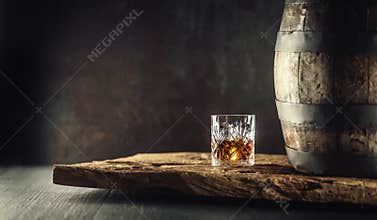 Glass of whisky or bourbon in ornamental glass next to a vinatge wooden barrel on a rustic wood and dark background