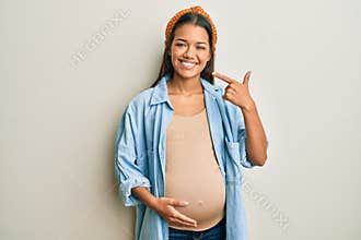 Beautiful hispanic woman expecting a baby, touching pregnant belly smiling cheerful showing and pointing with fingers teeth and