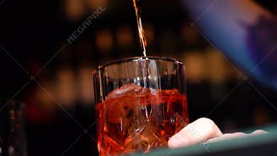 Barman pouring golden whiskey, cognac or brandy from bottle into glass with ice cubes on wooden table. Shiny gold party celebratio