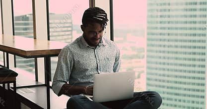 Happy young african american man working on computer, sitting on windowsill.